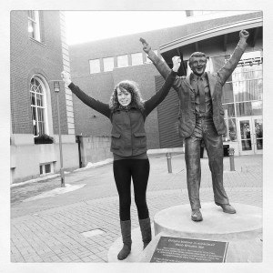 Maggie standing with arms raised next to Herb Brooks statue in Saint Paul, MN.
