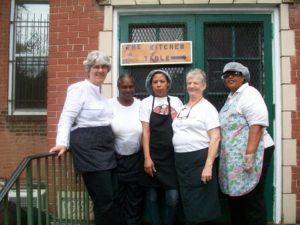 Photograph of five women, two with aprons nad hairnets, on steps in front of a sign that says "the kitchen table."