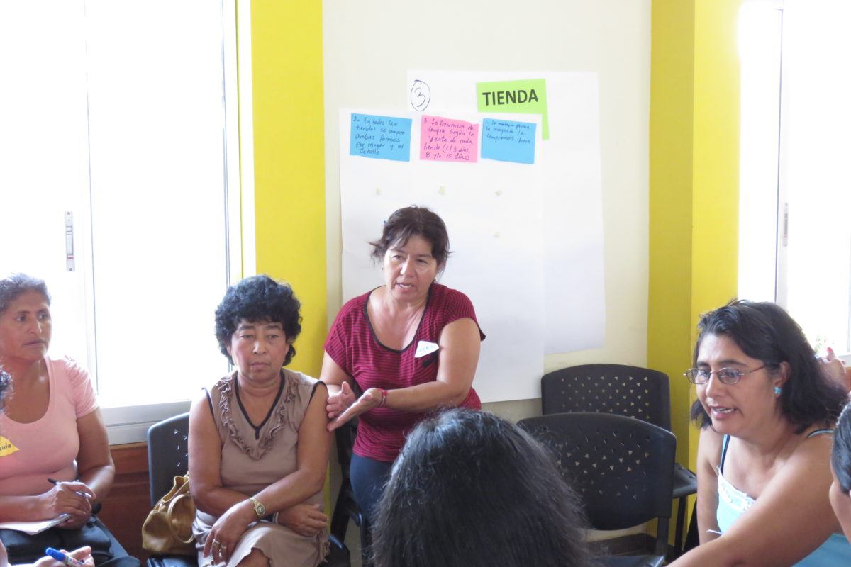 Photograph of a group of women sitting in a cirlce, one woman is talking. Behind her is flipchart paper that says tienda and has a list of ideas underneath it.