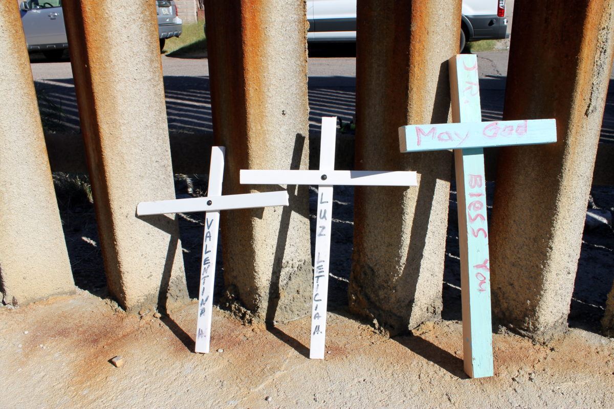 Photograph of three white crosses with names of victims on them leaning against the US/Mexico border.