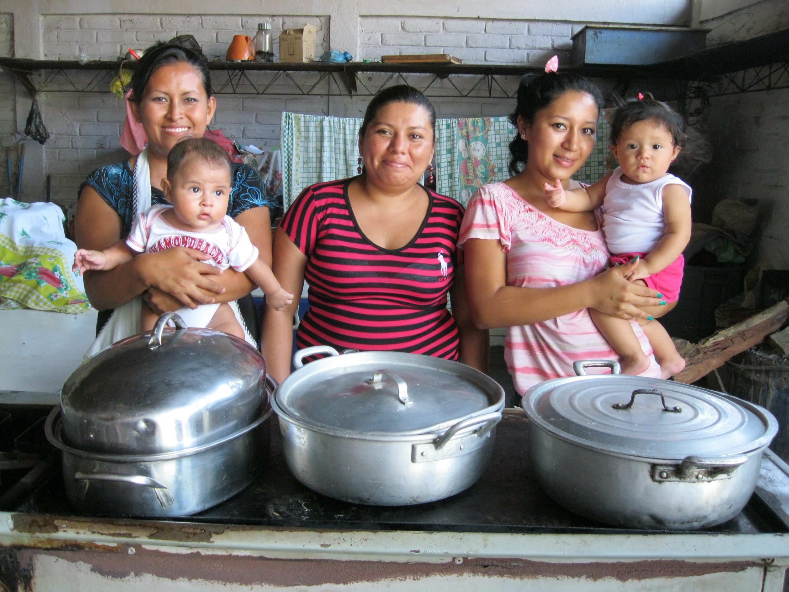 Photograph of three women, two of whom are holding babies, with three large cooking pots in front of them.