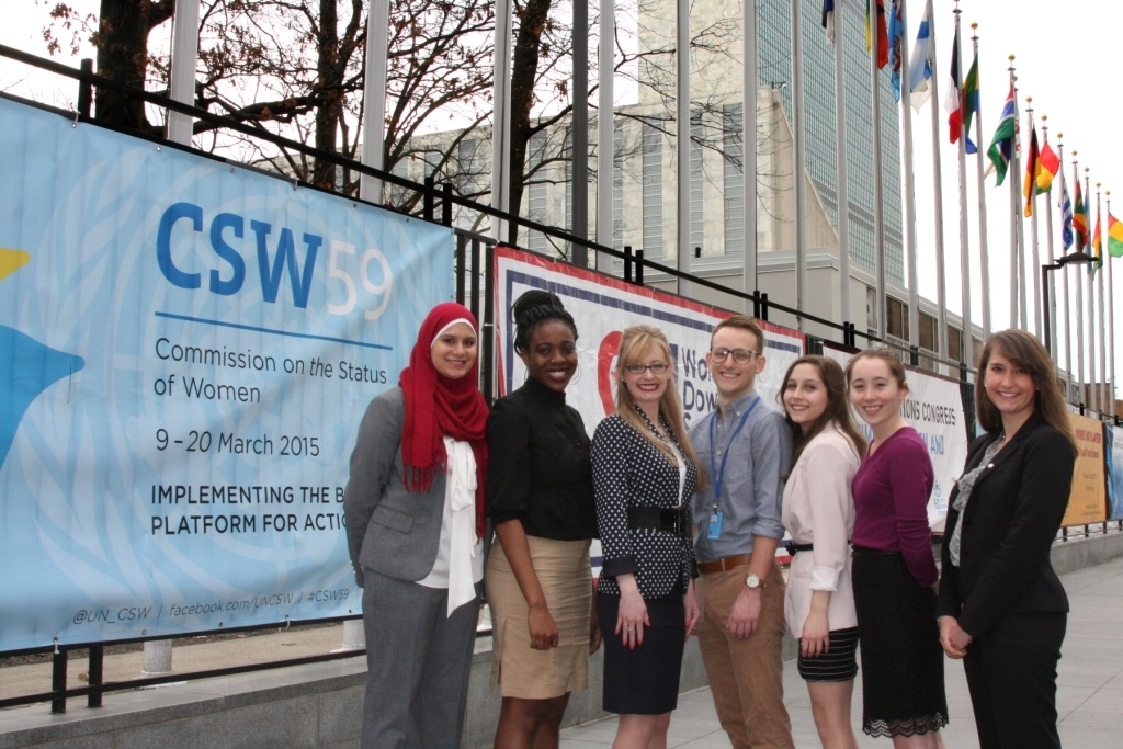 A group of six young women and one man stand in front of the United Nations with flags from various nationalities in the background and a poster that says CSW 59 Commission on the Status of Women