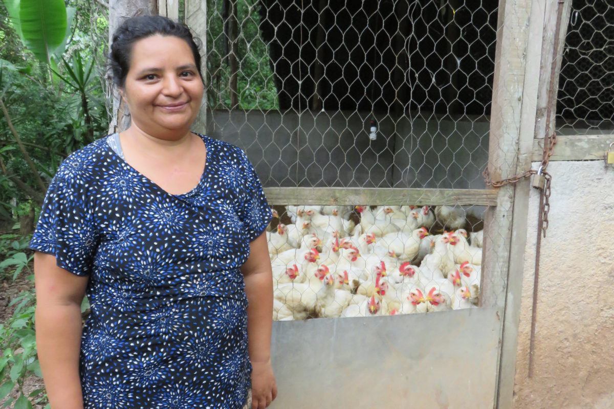 Isabel, a middle aged woman stands proudly in front of a chicken coop full of chickens.