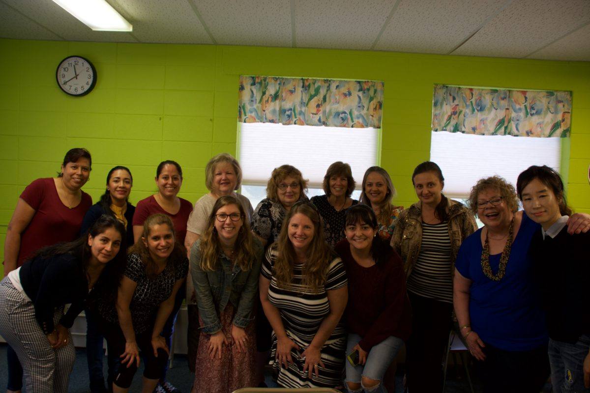 A diverse group of 15 women poses in two rows for the camera in a classroom.