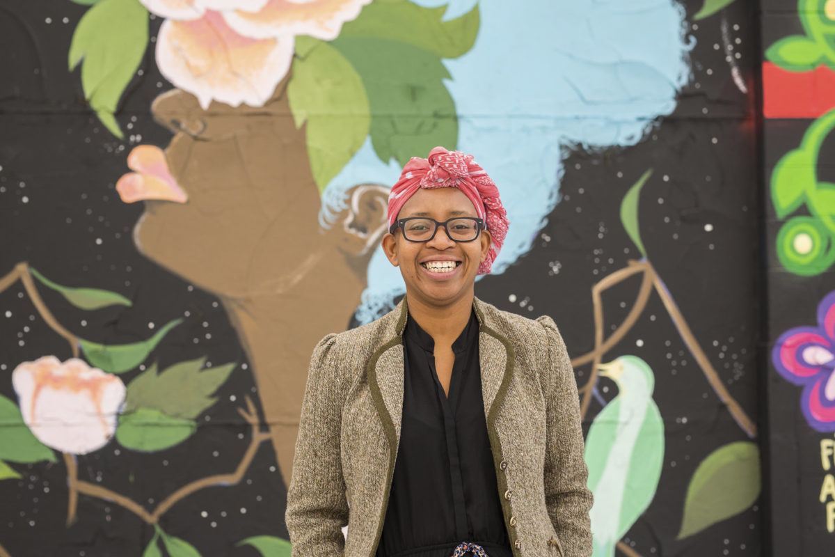 Photo of Nekessa Julia Opoti standing in front of a mural of the profile of a woman. Nekessa wears a red scarf on her head and glasses, and has a big smile.