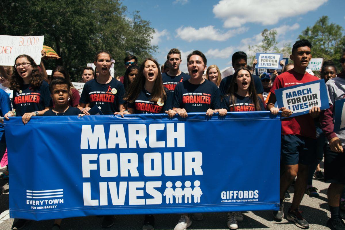 Photograph of many students marching outside and holding a blue sign reading "march for our lives"