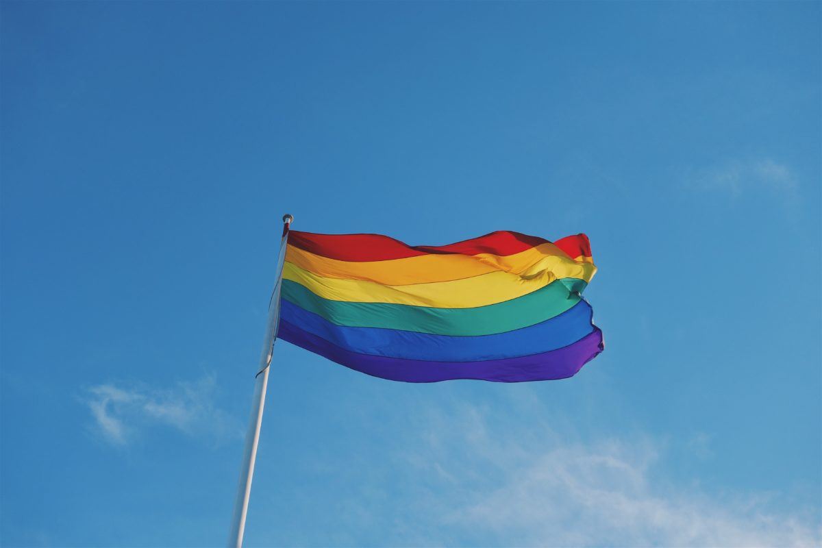 Photograph of LGBTQ+ Pride Flag waving against a clear blue sky.