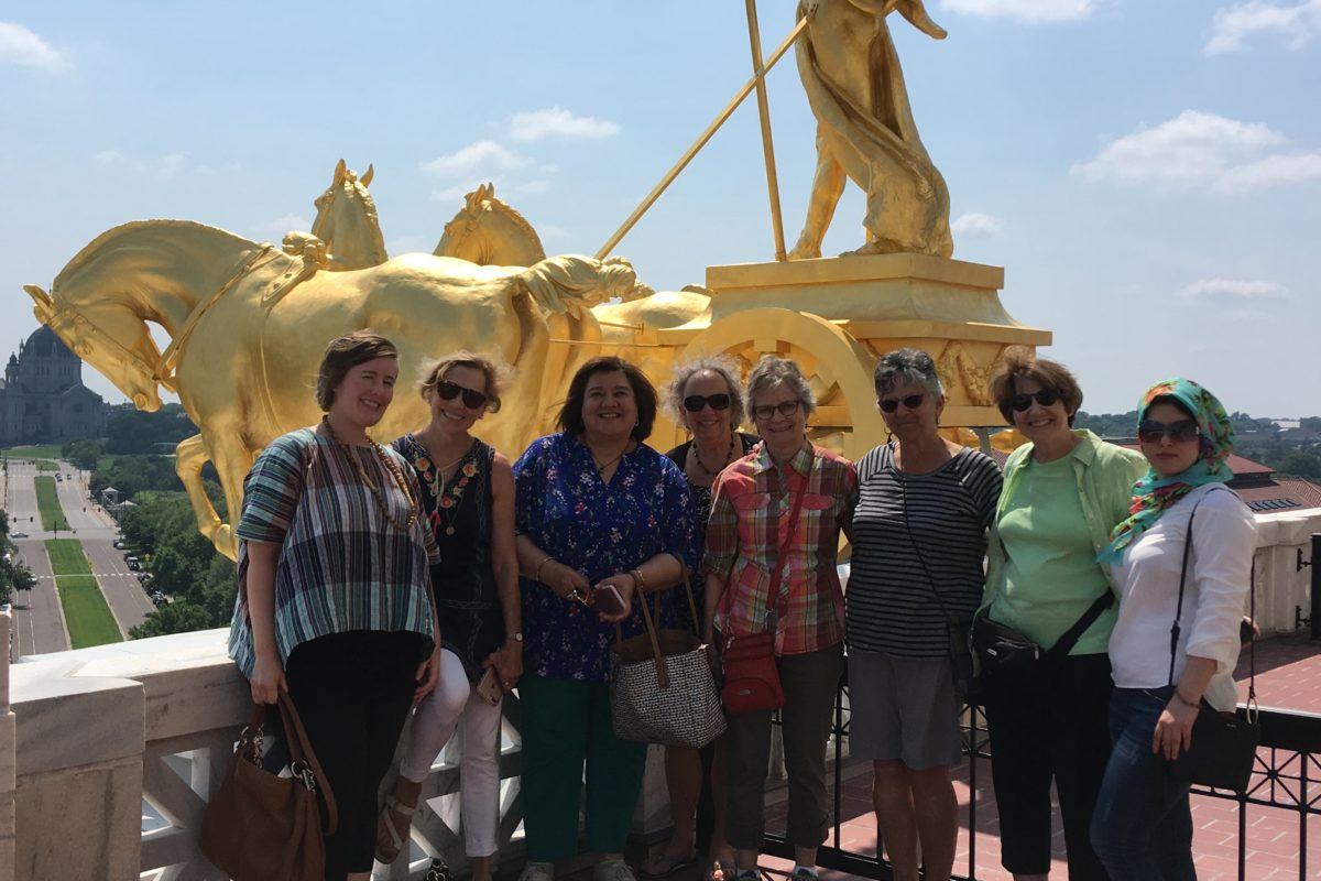 A group of seven women, some iraqi and some from the united states, stand in front of a golden statue on top of the Minnesota State Capitol building.